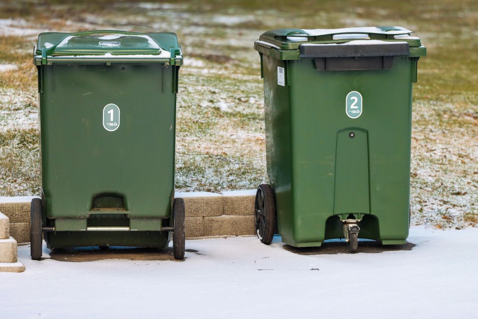 Two green wheeled plastic rubbish bins are positioned on a paved driveway with a low brick wall in front of a grassy area. The bin on the left has a flat, slightly textured lid with visible scratch marks and a label with the number '1' in a white circle. The bin on the right has a similar lid, but with a black hinge and a label displaying the number '2' in a white circle. Both bins are made of durable plastic with a smooth surface finish, and each has a pair of black wheels at the back, allowing for easier mobility. The ground below the bins appears to be dusted with snow, indicating cold weather, with some patches of snow visible on the driveway surface. The background includes grassy patches and a wooden or stone edging detail, suggesting a residential setting. This scene exemplifies typical private waste collection setup, highlighting independent rubbish bin management in a domestic environment under winter conditions. Waste Disposal Harringay provides rubbish removal services that support alternative waste handling outside local authority collection systems.