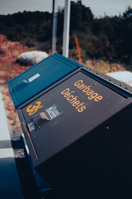 A large black waste disposal bin with a blue lid positioned on a paved surface, marked with bright orange text reading 'Garage Decants' and the number '36'. The container is made of durable plastic with a smooth finish and is situated outdoors, with a background showing a landscape of trees, some rocks, and a clear sky. The bin is partially tilted, indicating it might be in use or recently emptied, with sunlight casting shadows on its surface. The scene reflects a typical outdoor setting for private waste management or rubbish removal services, emphasizing the container's role in on-site clearance or alternative waste handling. The environment appears calm and functional, suitable for a professional rubbish collection service such as Waste Disposal Harringay, which caters to various waste disposal needs in the area.