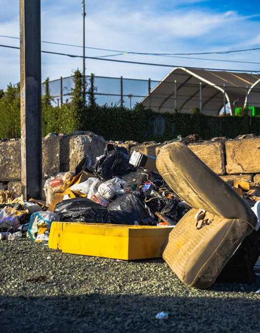 A roadside scene showing a pile of mixed rubbish including black and white plastic bags, discarded packaging, and a large, dirty, beige car seat placed atop the pile, with some debris spilling onto the gravel surface. Behind the rubbish, there is a low stone wall and a chain-link fence, with a hedge running along the fence line. In the background, a metal utility pole and overhead power lines are visible against a blue sky with some clouds. The scene appears to be an unauthorized or illegal rubbish dump, illustrating the importance of proper waste disposal and the services offered by Waste Disposal Harringay for prompt and responsible rubbish removal, especially when avoiding common mistakes in booking private or on-site clearance services.