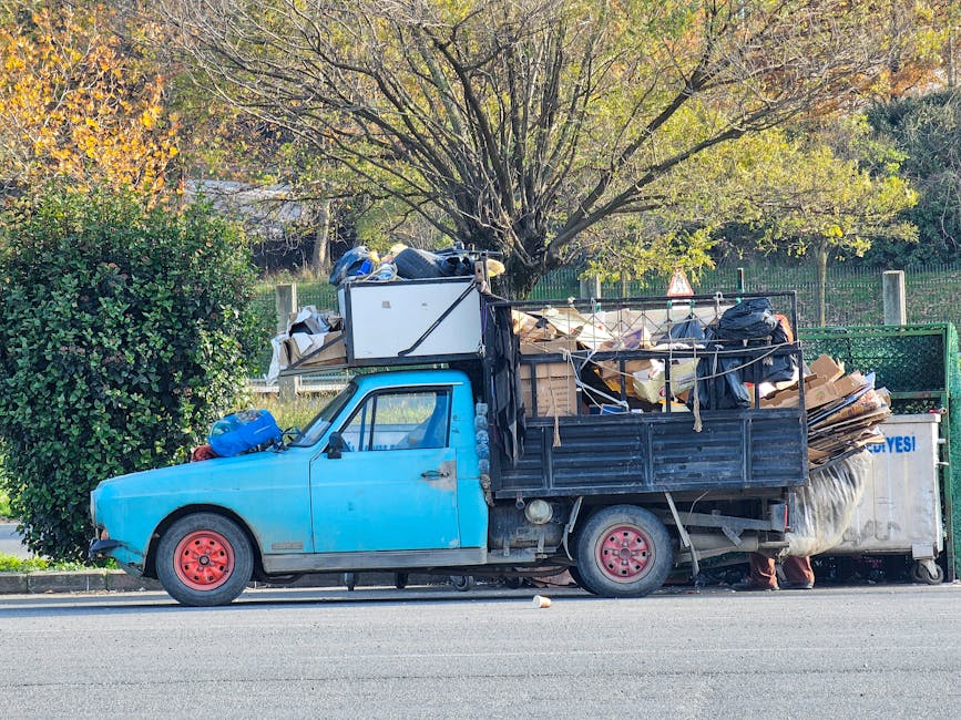 A blue flatbed truck parked on a street, heavily loaded with a mix of discarded cardboard boxes, black plastic bags, and various debris, with some items stacked and leaning against the truck's sides and rear. The truck has red wheels, and the load extends above the height of the vehicle, with some items secured by ropes or straps. In the background, there is a green hedge, a tall tree with branches and leaves displaying early autumn colors, and a metal fence. The scene is illuminated by natural daylight, highlighting the textures of the materials, including the rough surfaces of the cardboard and the smooth, shiny black plastic bags. The environment suggests an informal, private rubbish collection or on-site clearance, reflecting waste removal activity outside a typical municipal refuse service, which is relevant to the context of opting for alternative waste handling methods. Waste Disposal Harringay might use such trucks for efficient rubbish removal or private disposal services.