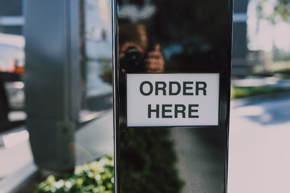 A black vertical post with a white rectangular sign attached, displaying the words 'ORDER HERE' in bold, uppercase black letters. The post is situated outdoors on a sidewalk or pavement area, with blurred background elements including greenery, a vehicle, and part of a parking lot. The lighting appears natural, suggesting daytime, and the reflective surface of the post hints at a metal or glossy finish. This sign is likely part of a drive-thru or outdoor ordering station, supporting private or independent waste management services such as waste disposal or rubbish collection near urban or commercial areas, aligning with the context of rubbish removal and alternative waste handling solutions by Waste Disposal Harringay.