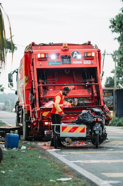 A waste collection worker, dressed in a red and yellow reflective uniform, is seen operating a large red rubbish collection truck on a roadside. The worker is standing at the rear of the vehicle, emptying waste into the hopper, with their hands engaged in the task. The back of the truck displays safety markings, a license plate number 'BRJ 775A', and various warning labels. In front of the truck, a small motorbike is parked, with a black plastic trash bag placed on its seat, adjacent to a metal guardrail. The environment includes a paved road with a painted curb, and in the background, there are trees and a commercial building with signage, hinting at an urban or suburban area. Surrounding the scene is a grassy verge with some scattered debris and a blue container visible near the roadside. The lighting suggests daylight conditions, and the scene captures an on-site waste collection operation, reflecting an alternative waste handling activity that might be part of a private rubbish removal service, as indicated by the presence of Waste Disposal Harringay in the broader context.