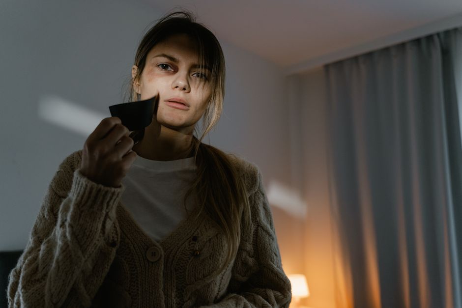 A young woman with long brown hair and light skin is holding a black remote control in her right hand, positioning it closer to the camera while looking directly ahead with a neutral or slightly puzzled expression. She is wearing a beige knitted cardigan over a white top. The background features a softly lit indoor environment with a warm-toned lamp on a side table, and a large window with pale blue or gray curtains that are partly drawn, allowing some natural light into the room. The setting appears to be a living room or a lounge area, with the focus on her upper body and face. The scene’s lighting creates gentle shadows, emphasizing her facial features and the texture of her clothing. The image suggests a scenario where the woman might be interacting with electronic devices or contemplating a decision, fitting within a context of household or everyday activities related to waste disposal or general home management, subtly aligning with the themes of independent waste handling or on-site clearance provided by Waste Disposal Harringay.