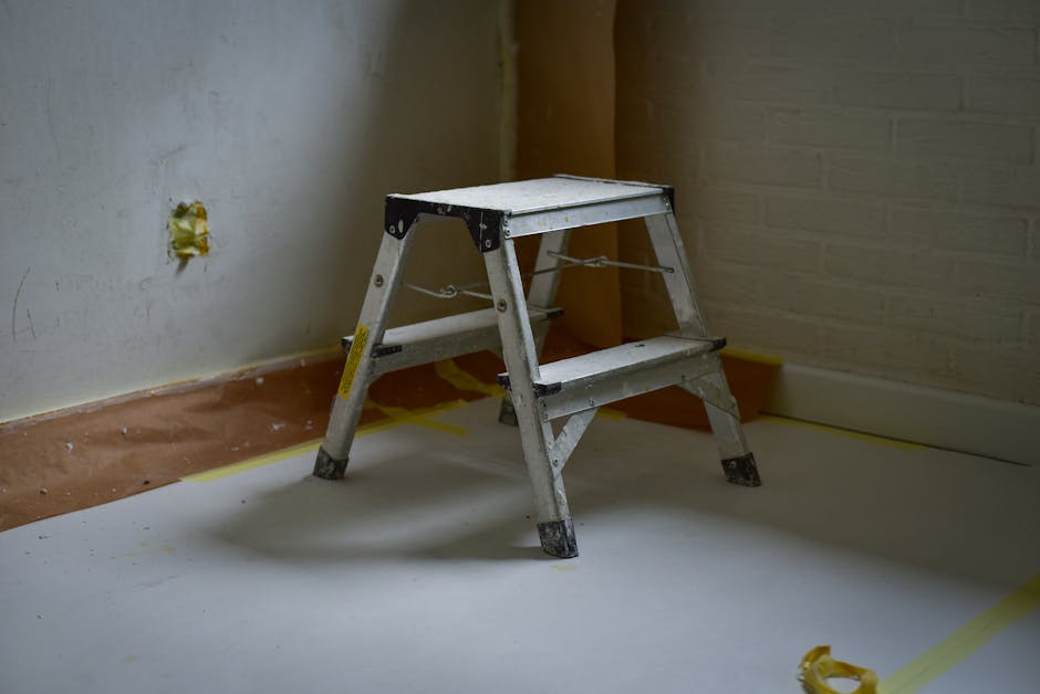 A silver metal step ladder with black plastic feet is positioned in the middle of a room under renovation. The ladder has two steps and appears slightly worn, with some visible scratches and dirt on its surface. It is placed on a partially covered floor, which is protected by a white sheet or plastic sheeting, secured at the edges with yellow painter's tape. Behind the ladder, a white brick wall is visible on the right, and a plain, light-colored wall with an unfinished electrical outlet on the left, where some yellow insulation or filler is exposed around the socket. The environment suggests an indoor space undergoing refurbishment or decoration, with some construction debris or marks on the walls and floor. Waste Disposal Harringay may be involved in clearing or removing refuse associated with such renovation activities, reflecting the company's scope of services in private or independent waste collection for property renovation or end-of-tenancy clearance scenarios.