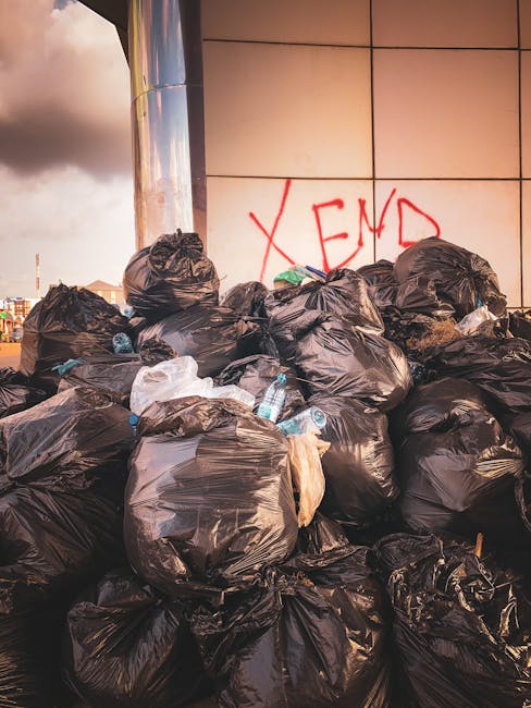 A large pile of black plastic garbage bags filled with rubbish is stacked against a building wall with a tiled surface. Some bags are torn open, revealing discarded plastic bottles and other waste material inside. The wall features red graffiti text, partially visible as 'XEND'. The scene is outdoors, with natural lighting casting shadows on the bags and wall, suggesting late afternoon or early evening. The surrounding environment appears to be a city or urban area, possibly near a commercial or residential building, indicating a situation of unmanaged waste that might require private rubbish removal services like those offered by Waste Disposal Harringay for end-of-tenancy or on-site clearance purposes.