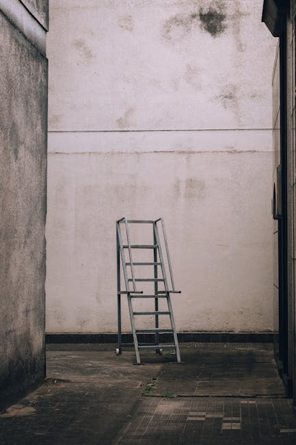 A silver metal step ladder with black plastic feet is positioned in the middle of a room under renovation. The ladder has two steps and appears slightly worn, with some visible scratches and dirt on its surface. It is placed on a partially covered floor, which is protected by a white sheet or plastic sheeting, secured at the edges with yellow painter's tape. Behind the ladder, a white brick wall is visible on the right, and a plain, light-colored wall with an unfinished electrical outlet on the left, where some yellow insulation or filler is exposed around the socket. The environment suggests an indoor space undergoing refurbishment or decoration, with some construction debris or marks on the walls and floor. Waste Disposal Harringay may be involved in clearing or removing refuse associated with such renovation activities, reflecting the company's scope of services in private or independent waste collection for property renovation or end-of-tenancy clearance scenarios.