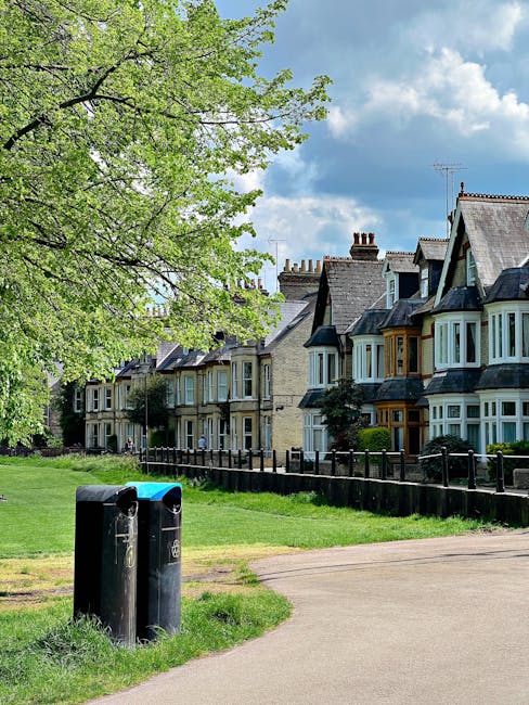 A row of Victorian terraced houses with varying architectural details, including bay windows and pitched roofs, situated along a residential street. In the foreground, two black waste disposal bins are positioned on a paved path next to a grassy verge. To the left, a large, leafy tree with light green foliage extends its branches overhead, partially shading the scene. The background features a bright blue sky with scattered white clouds, and the scene appears well-maintained and tidy. The environment suggests a peaceful urban neighbourhood that may benefit from private rubbish collection services, such as those offered by Waste Disposal Harringay, for efficient waste management engagement.