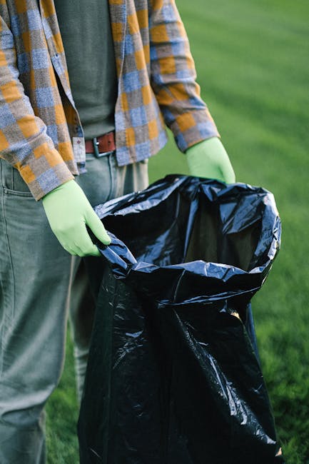 A person wearing a yellow and gray plaid shirt, green gloves, and gray pants is holding open a large black plastic rubbish bag outdoors on a grassy area. The bag appears to be made of shiny, durable plastic material suitable for waste disposal. In the background, there is a blurred expanse of green grass, indicating a park or garden setting. The scene suggests they are preparing to fill the bag with rubbish, possibly related to garden or outdoor waste collection, providing a visual example of private waste handling or on-site clearance services offered by Waste Disposal Harringay. The lighting is natural, with soft daylight illuminating the scene, emphasizing the texture of the rubbish bag and the fabric of the person's clothing. The image captures a moment of waste disposal activity, aligned with the theme of rubbish removal and alternative waste management methods outside the context of municipal collection services.