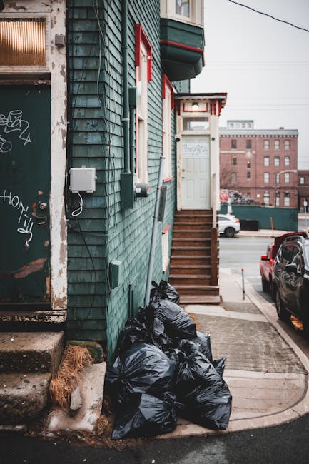 A photograph captures the exterior of a narrow residential building with green wooden cladding showing signs of weathering. Several black rubbish bags, filled and tied, are stacked at the base of the building near a concrete step. The bags are placed beside an aged, graffiti-covered door with a rusted frame, which appears to be part of a secondary entrance or utility area. A small, white outdoor electrical box is mounted on the wall nearby, with a cable running from it. Wooden stairs with a simple handrail lead up to a white door framed by a small porch, indicating a private entrance. In the background, a city street scene is visible featuring parked cars, including a black vehicle closest to the building, and a distant red-brick industrial building with multiple windows. The scene is illuminated by natural daylight, creating an everyday urban atmosphere. The image relates to rubbish removal services, illustrating an instance of private waste disposal, possibly involving independent collection or on-site clearance managed by Waste Disposal Harringay, serving properties with limited access.