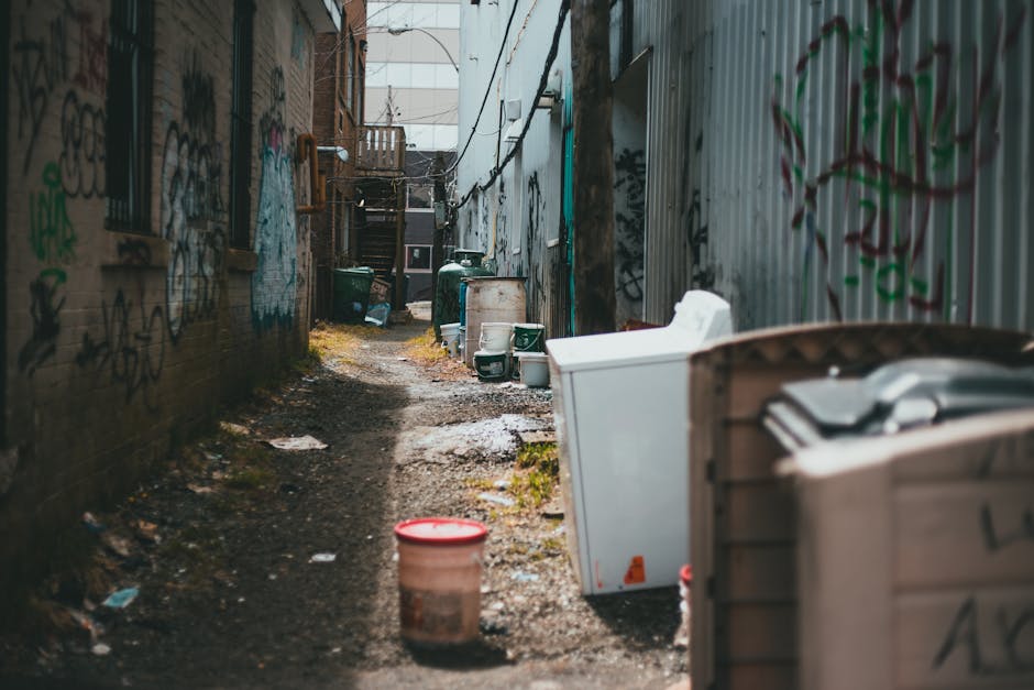The image depicts a narrow alleyway between two buildings with graffiti-covered brick and corrugated metal walls on either side. The ground is uneven, muddy, and strewn with small debris, indicating a lack of regular maintenance. Several large waste containers, including a white plastic skip and green wheelie bins, are positioned along the alley, with some lids open and debris visible nearby, suggesting ongoing rubbish removal or disposal activities. In the foreground, a red and beige plastic bucket sits on the ground next to the containers. The alley extends toward a set of external wooden stairs at the far end, with additional waste bins visible further back. Overhead, electrical wires crisscross the space, and some natural daylight filters in from above, illuminating the scene with a neutral tone. The environment reflects an area that may benefit from professional waste management services, such as those provided by Waste Disposal Harringay, targeted at managing rubbish in spaces with limited access to municipal waste collection.