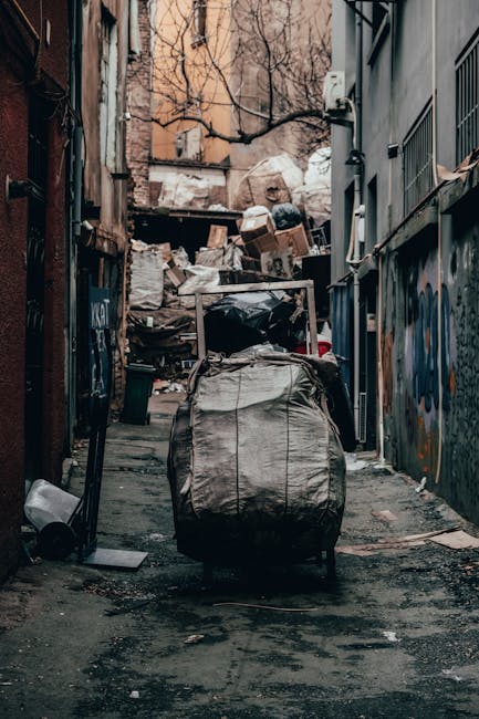 A narrow urban alleyway filled with a large, wheeled trash bag in the foreground, made of heavy-duty, weathered grey plastic with visible creases and dirt marks. Behind it, there are overflowing cardboard boxes, plastic bags, and discarded debris piled against a makeshift barrier of wooden pallets and other materials at the far end of the alley. The surrounding walls are constructed from brick and metal, showing signs of graffiti and weathering, with some graffiti visible on the right wall. The ground is uneven, stained with dark patches, and scattered with small bits of litter. Bare tree branches are visible overhead, indicating an outdoor setting in a city environment. The scene is illuminated by natural diffused light, suggesting an overcast day. This scene exemplifies an area where private waste collection or rubbish removal services, such as those provided by Waste Disposal Harringay, are often employed to manage urban debris in alleys and backstreets requiring urgent rubbish clearance.