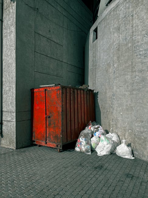 A rusty red metal skip positioned against a dark green wall and a light gray concrete wall on a paved urban street or alleyway, with several clear plastic rubbish bags containing assorted waste such as plastic bottles and packaging stacked on the ground in front of it. The skip appears old and weathered, with visible patches of rust and peeling paint, while the plastic rubbish bags are loosely tied and slightly crumpled, indicating they are awaiting collection or disposal. The surrounding environment suggests an outdoor setting in a city or town area, with no visible signage or vehicle nearby. The scene reflects an example of private waste storage or handling, often associated with independent rubbish removal services, as seen by Waste Disposal Harringay's service focus on alternative waste management solutions without relying on municipal collection. Lighting is natural, casting soft shadows on the ground, highlighting the textures of the skip, bags, and pavement, and emphasizing the utilitarian nature of this urban waste disposal scene.